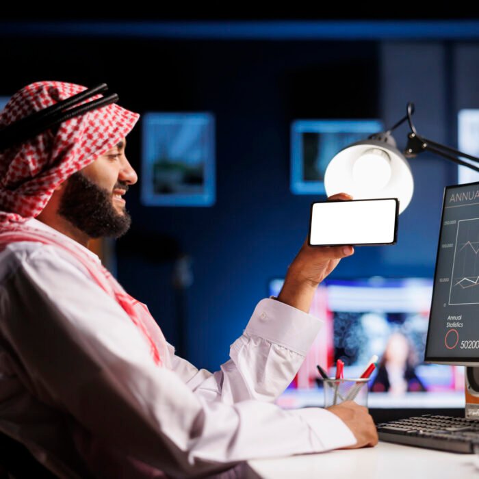 Mobile phone displays white screen An Arabic individual, wearing traditional clothing uses a mobile phone at an office desk. The isolated white screen offers space for customized content in an advertising or promotional setting.